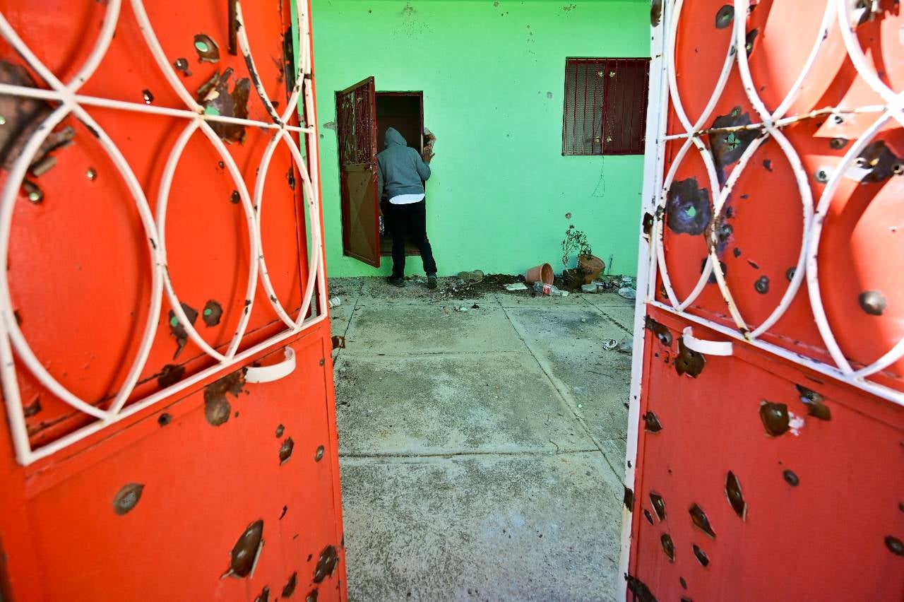 Un hombre ingresa a una casa con agujeros de bala en el portón, que fue saqueada por miembros de una organización criminal.